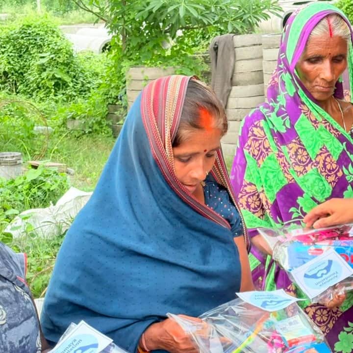 Two women in saris looking down at transparent packs of sanitary pads and toiletries labelled Annmaya Foundation, held in their hands on open ground