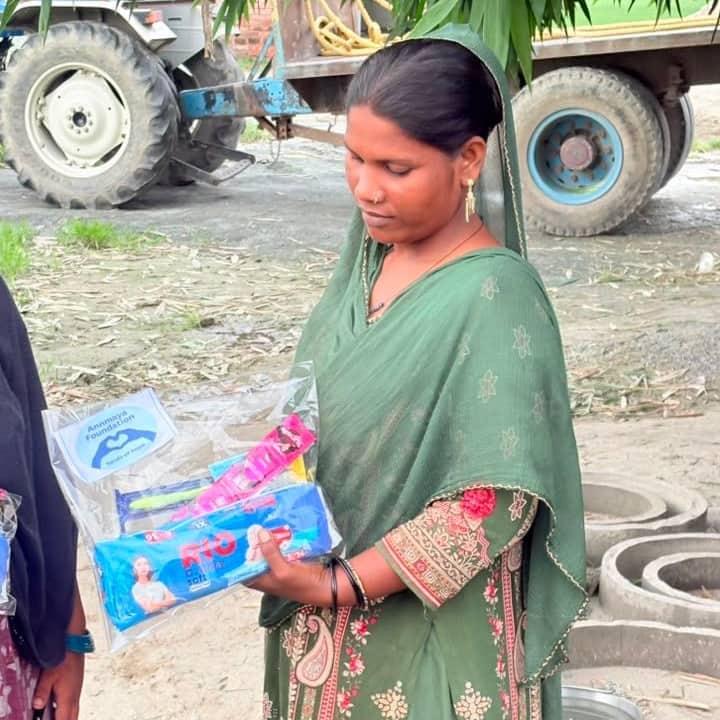 A woman in a green sari holding a transparent pack of sanitary pads and toiletries labelled Annmaya Foundation, with a tractor parked behind her