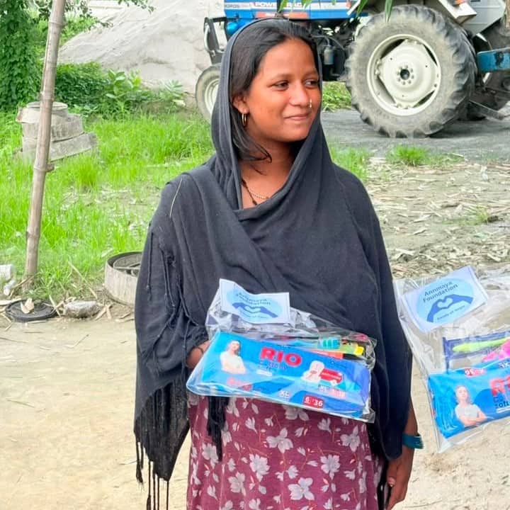 A woman in a dark headscarf holding two transparent packs of sanitary pads and toiletries labelled Annmaya Foundation, standing on open ground beside a tractor