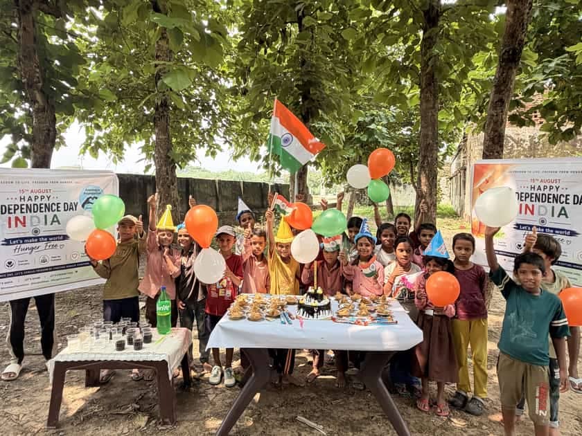 Children in paper party hats holding orange, white and green balloons and an Indian flag behind a table laid with a cake and plates of food, under trees, beside Annmaya Foundation’s 15 August banner